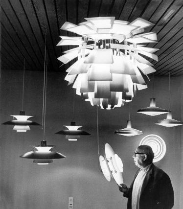 Een historische zwart-witfoto van Poul Henningsen met zijn iconische lampen voor Louis Poulsen. Op de voorgrond houdt Henningsen een lampmodel in zijn hand, terwijl boven hem verschillende van zijn ontwerpen hangen – waaronder de beroemde Artichoke-lamp, die met zijn vele lamellen een zacht, niet-verblindend licht produceert.