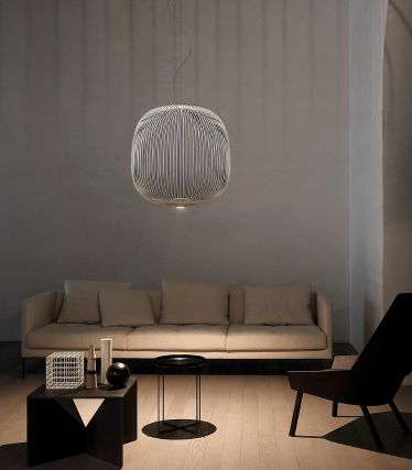Een minimalistische woonkamer met de Spokes hanglamp van Foscarini als elegant middelpunt. De filigrane, metalen steunen van de lamp werpen zachte lichtspelen op de muur en het plafond en geven de ruimte een licht, sculpturaal effect. Een lichte bank, donkere accenten en natuurlijke materialen creëren een rustige, moderne sfeer.