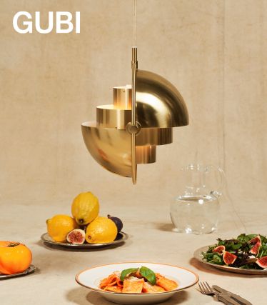 Een elegante Multi-Lite pendellamp van GUBI in geborsteld messing hangt boven een gedekte tafel. Daaronder staan borden met pasta, verse vijgen en citroenen en een glazen karaf – een stijlvol tafereel dat design en genot combineert.