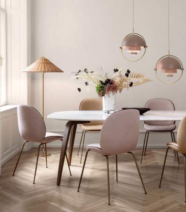 Een zachte, elegante eetkamer met een witte marmeren tafel en pastelroze en beige stoelen op messing poten. Boven de tafel hangen twee ronde hanglampen in warme beige tinten met messing details. Bij het raam staat een hoge staande lamp met een geweven kap en op de tafel staat een vaas met gedroogde bloemen. De kamer heeft een lichte visgraatparketvloer en een rustig, neutraal kleurenpalet.