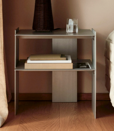 Smal metalen nachtkastje met twee planken, boeken, een glas en een vaas naast een bed.