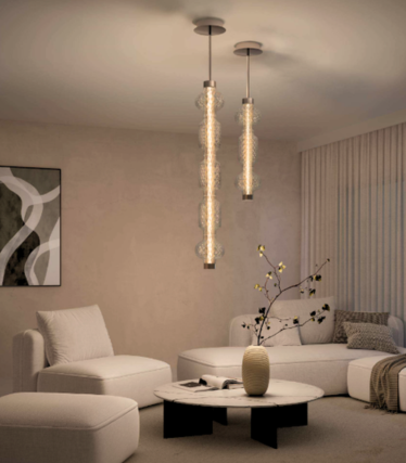 Twee slanke glazen hanglampen boven een marmeren salontafel in een lichte woonkamer met een witte bank en fauteuil.