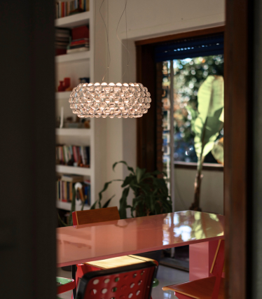 Bolvormige hanglamp van glazen bollen boven een rode eettafel, gezien door een deuropening.