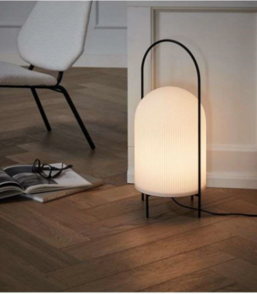 Witte geribbelde lamp met zwart beugelframe op visgraatparket naast een fauteuil.
