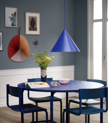 Blauwe kegelvormige hanglamp boven een ronde blauwe eettafel met een boeket bloemen, in een eetkamer met een blauwe wand en kunstwerken.