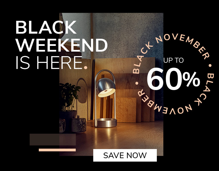 Modern silver table lamp glowing on a wooden desk with cutting boards and plants in the background. Promotional text reads: ‘Black Weekend is here. Up to 60%. Black November.’ A button says ‘Save Now.’